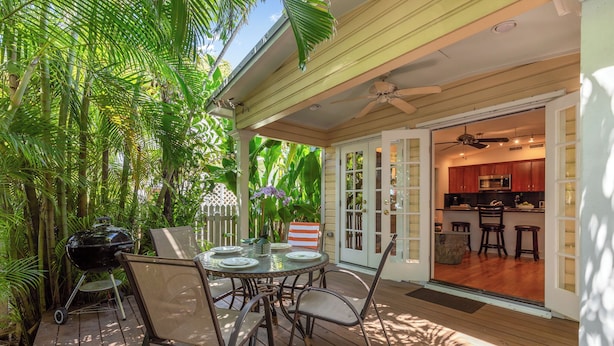 A wooden deck with a table and chairs, a grill, and a view into a kitchen through glass doors.