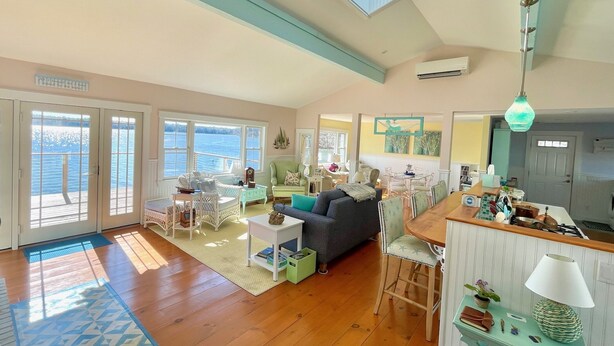 A spacious living room with a high ceiling, large windows, and a skylight. There is a dining area with a wooden table and chairs, and a kitchen area visible in the background. - Maine Waterfront Cottage: Nestled on an acre of land, with 500ft of shore front (Newcastle)
