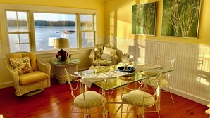 A room with a glass table surrounded by chairs, a wicker armchair, and a sofa. Large windows offer a view of a body of water. - Maine Waterfront Cottage: Nestled on an acre of land, with 500ft of shore front (Newcastle)