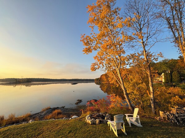 A lakeside view with chairs and a fire pit, surrounded by autumn foliage.