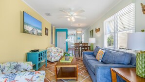 A living room with a blue sofa, a wooden coffee table, and a colorful chair. There is a dining area in the background with a table and chairs. - THE QUARTERDECK ~ Colorful & Comfy 2nd Floor 2Ba/ 1Ba Shipyard Unit! (Key West)