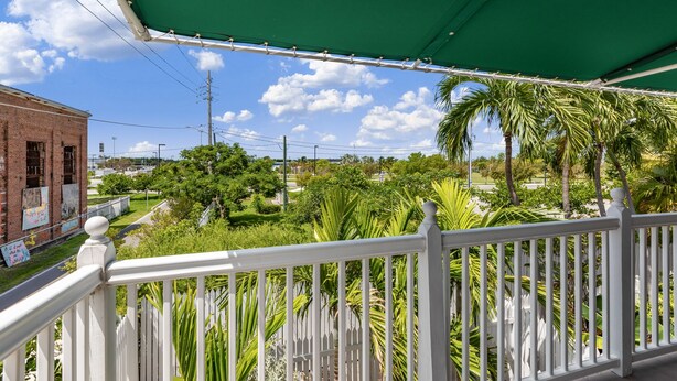 A balcony with a white railing overlooking a lush garden with palm trees and a brick building in the background. - THE QUARTERDECK ~ Colorful & Comfy 2nd Floor 2Ba/ 1Ba Shipyard Unit! (Key West)