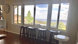 A living room with a wooden floor, a sofa, and three bar stools. Large windows offer a view of the outdoors. - Mt Solitary House – Elite Views | Incredible Comfort (Katoomba)