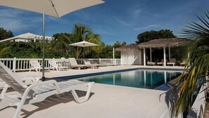 Piscina con sillas de descanso y sombrillas, rodeada de palmeras y una estructura de techo de paja.