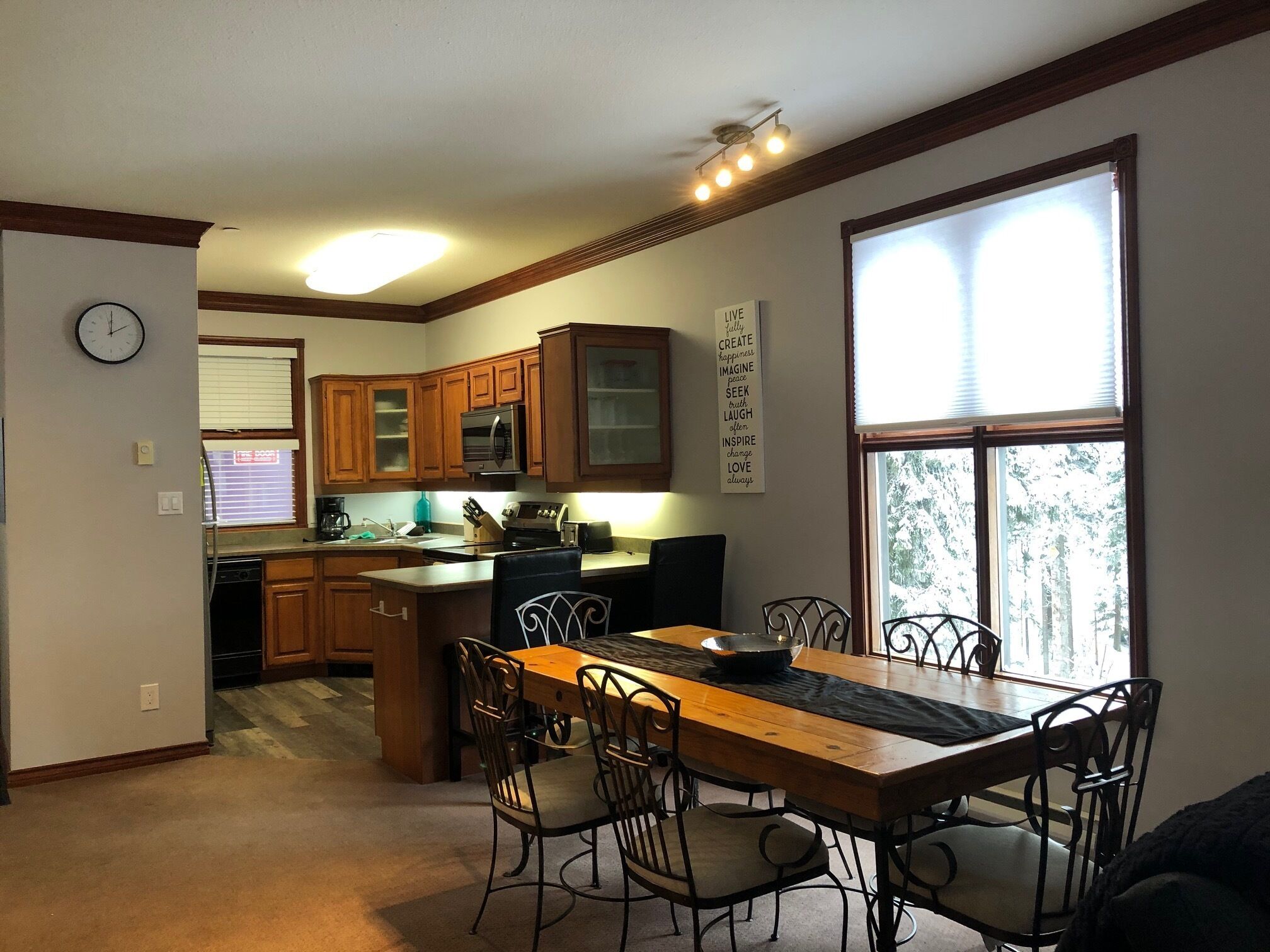 A kitchen with wooden cabinets, a dining table with chairs, a clock, and a motivational poster.