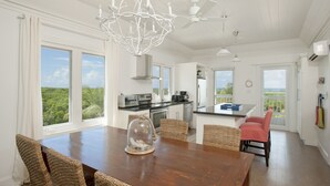A spacious kitchen with large windows, a dining area with wicker chairs, and a view of the outdoors. - AS SEEN ON HGTV!  Lux Condo w/Views, Htd Pool, Beach, Golf Cart, Pickleball (Governors Harbour)