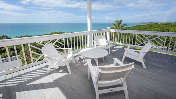 A wooden deck with white chairs and a table, overlooking a clear blue sea and lush greenery. - AS SEEN ON HGTV!  Lux Condo w/Views, Htd Pool, Beach, Golf Cart, Pickleball (Governors Harbour)