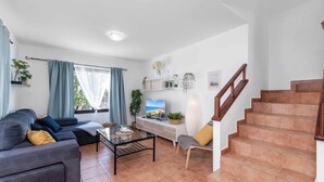 A modern living room with a sofa, coffee table, and a TV. There is a staircase with wooden railings and a view of the outdoors.