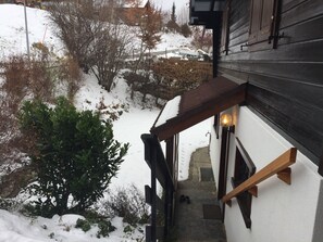 Escada de madeira que leva a um edifício com o chão coberto de neve e uma pequena árvore ao lado.