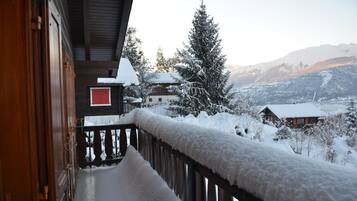 Ein Holzbalkon mit schneebedeckten Geländern und Blick auf schneebedeckte Häuser und Bäume in einer Berglandschaft.