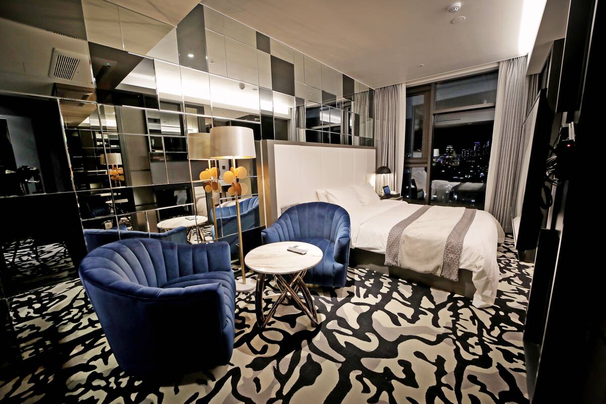 A modern hotel room with a large bed, two blue armchairs, a small round table, and a city view through the window.