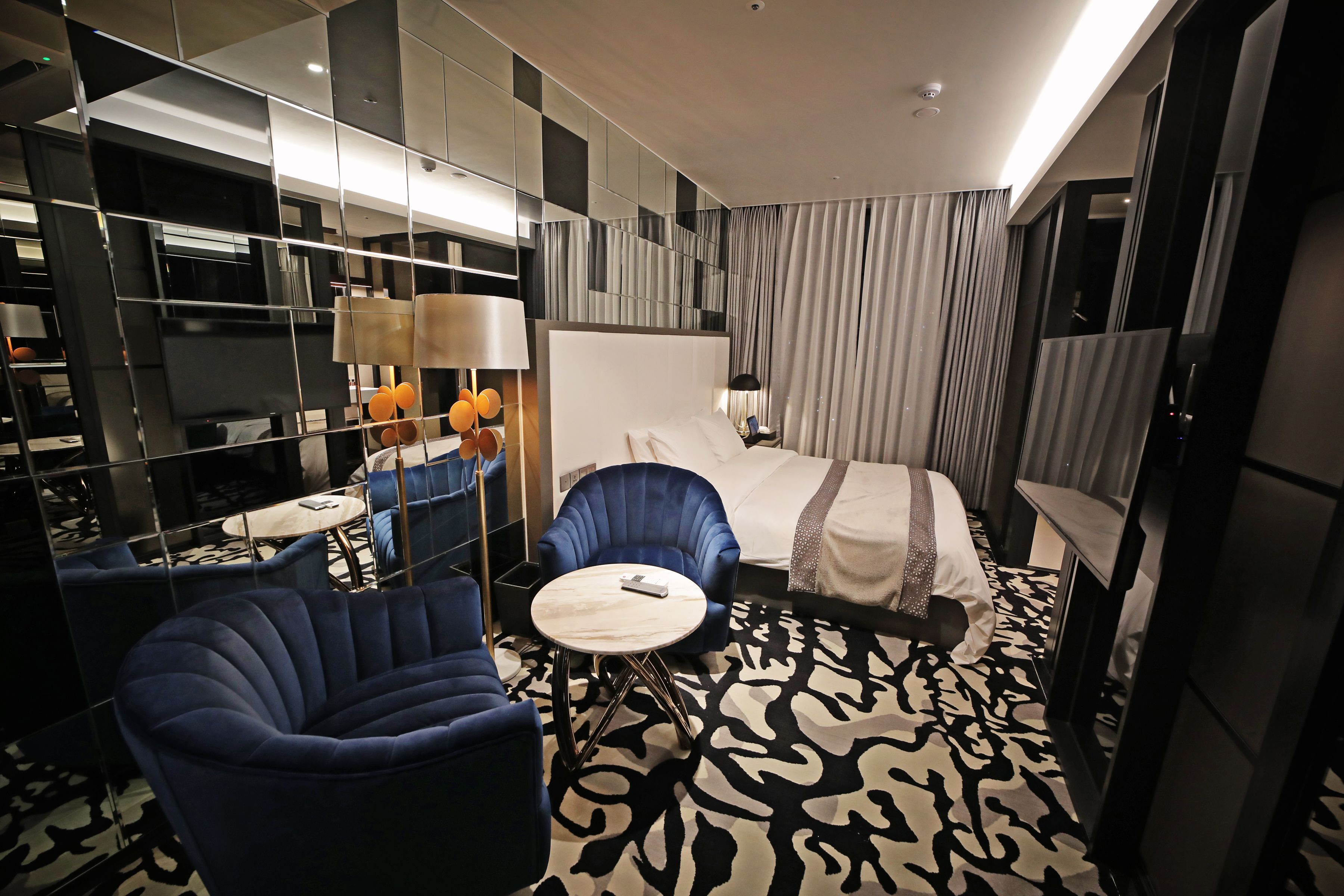 A modern hotel room with a large bed, a blue armchair, a small round table, and a large mirror.
