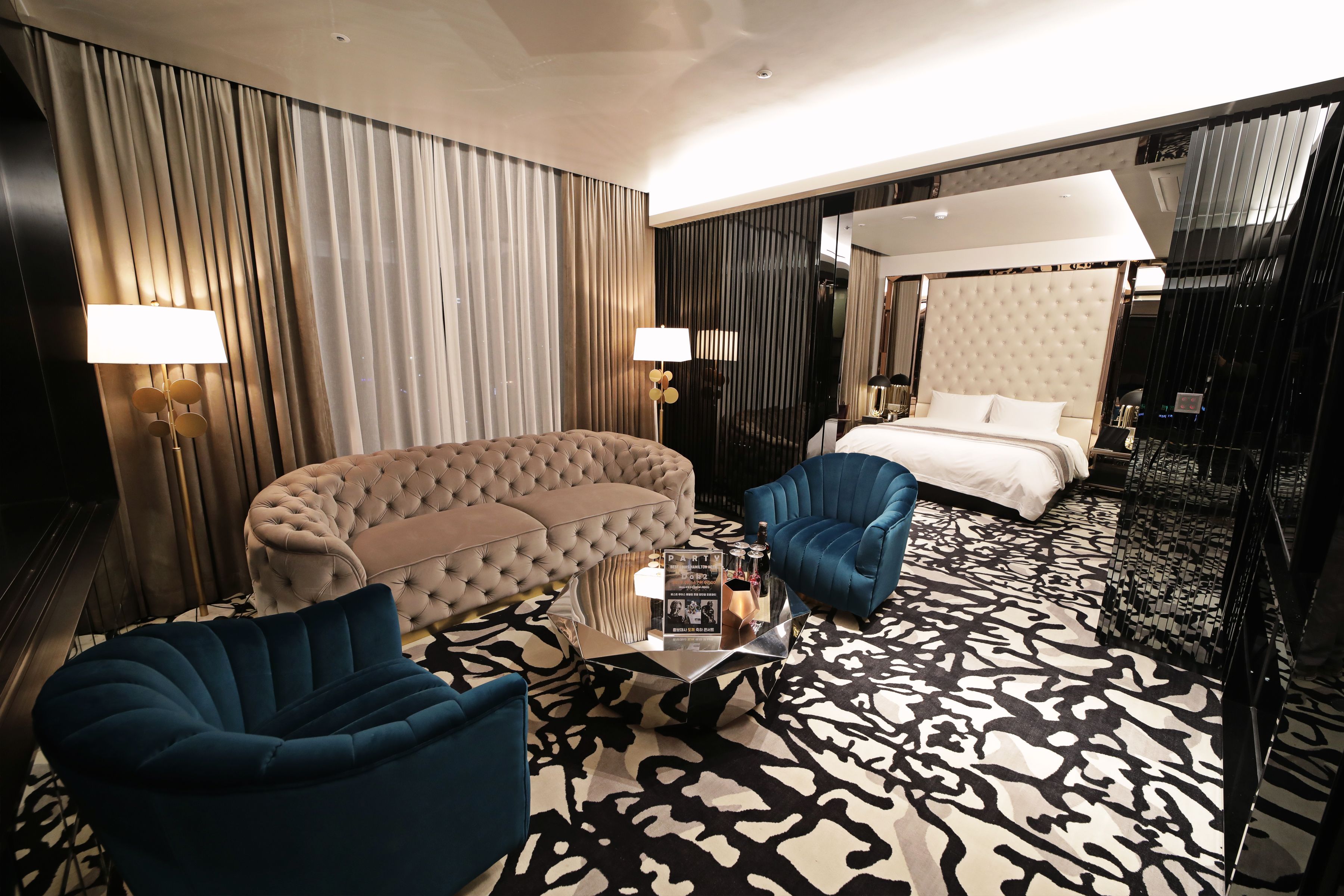 Ein modernes Hotelzimmer mit einer gesteppten Couch, einem blauen Sessel, einem Glastisch und einem gemusterten Teppich.