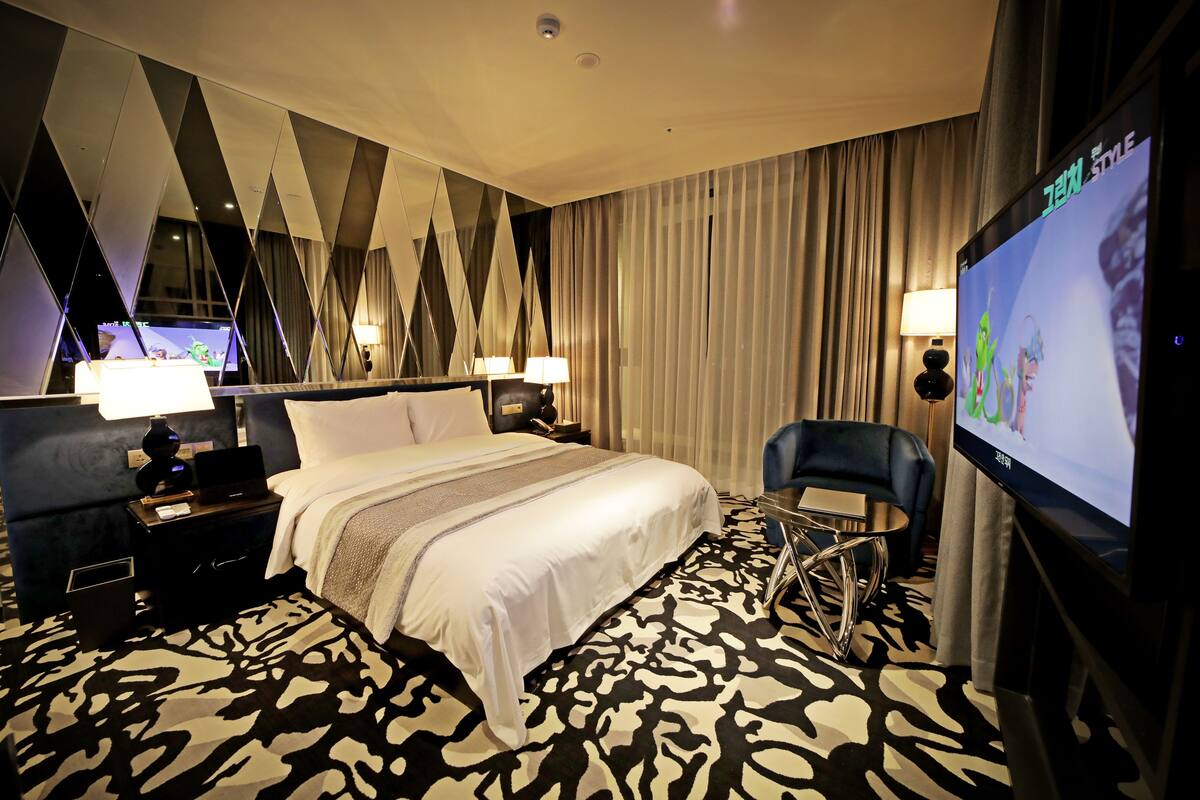 A hotel room with a large bed, two bedside tables with lamps, a TV, a chair, and a patterned carpet.