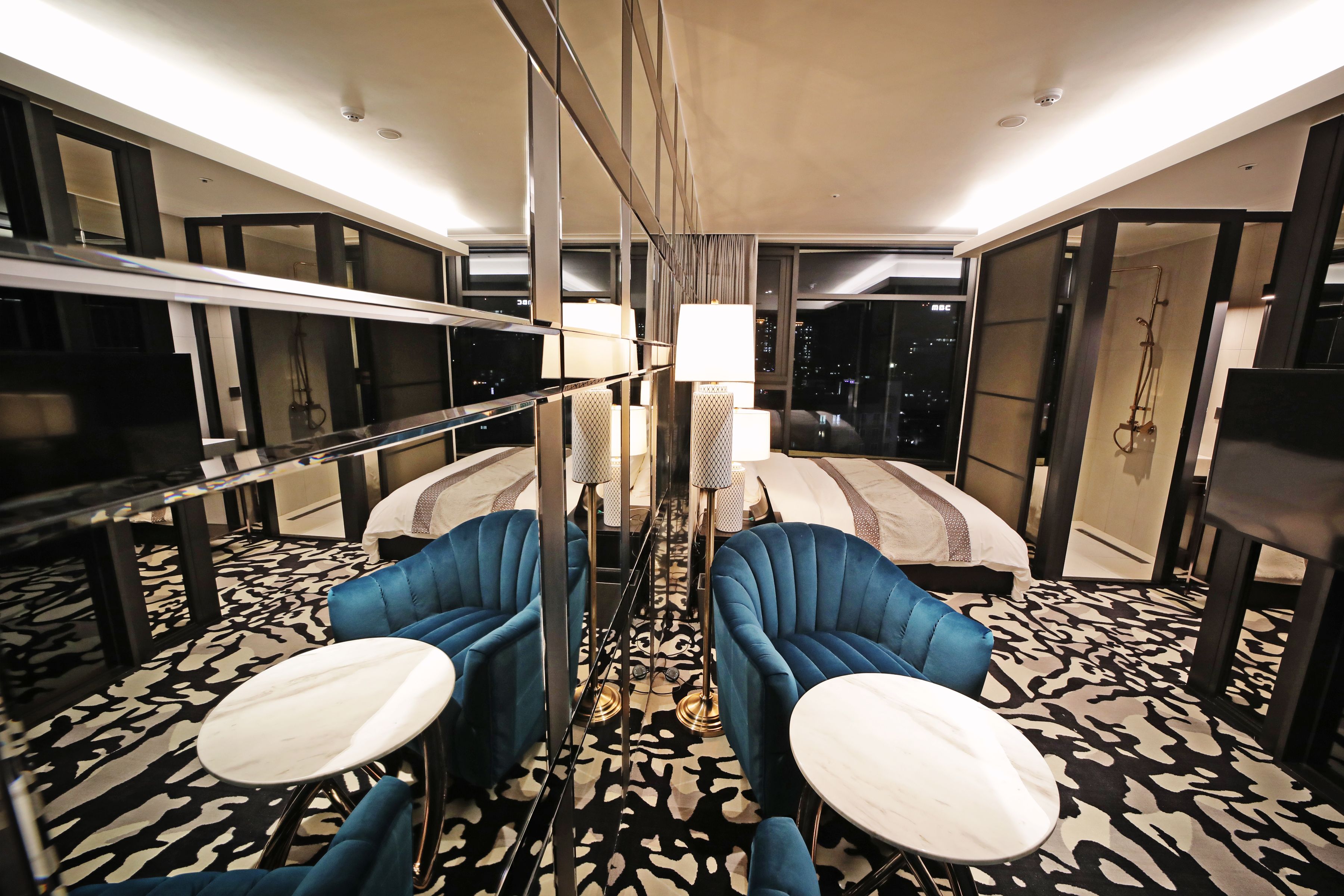A hotel room with a bed, two blue armchairs, a small table, and a large mirror.