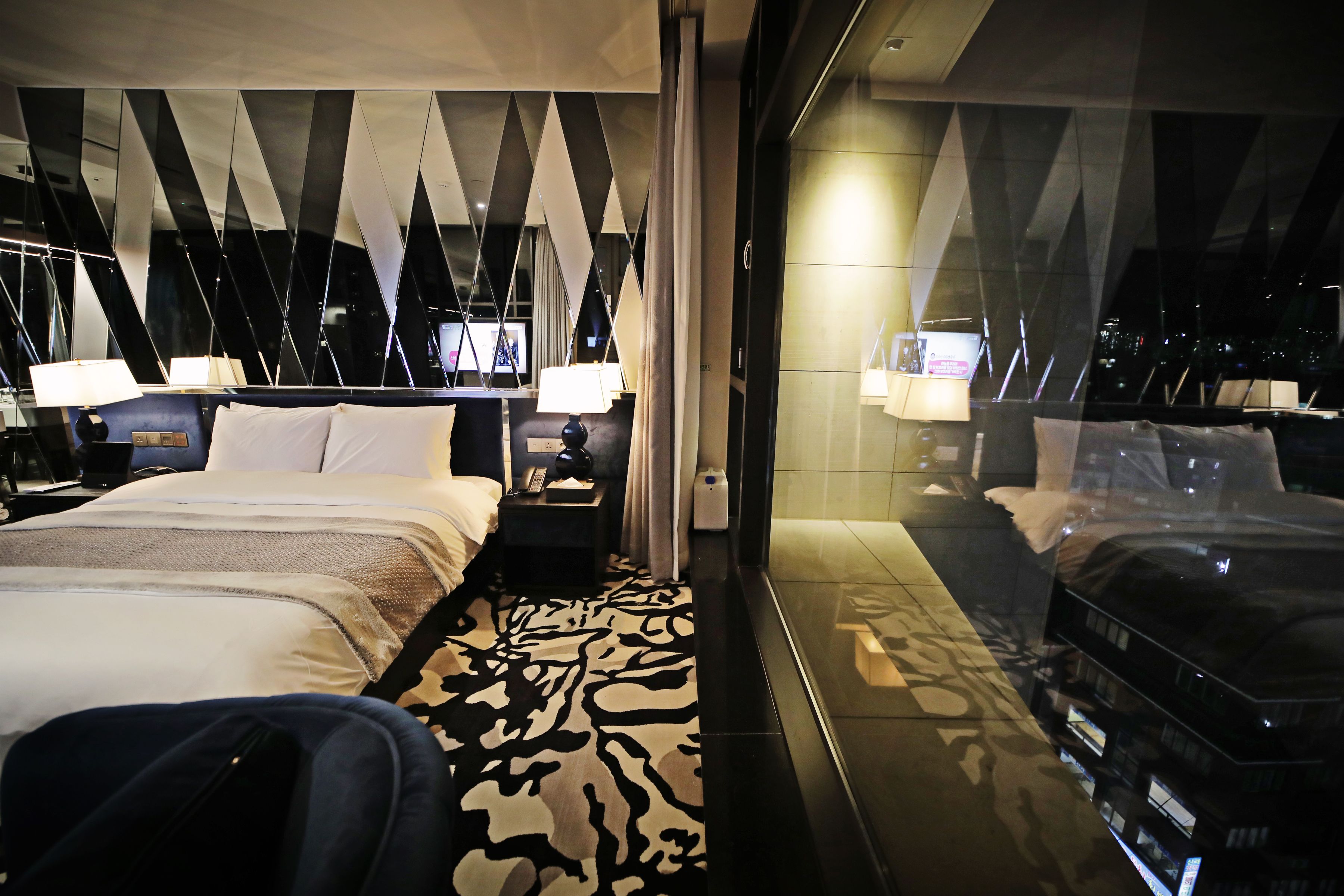 A hotel room with a bed, a patterned rug, a suitcase, and a large window with a city view.