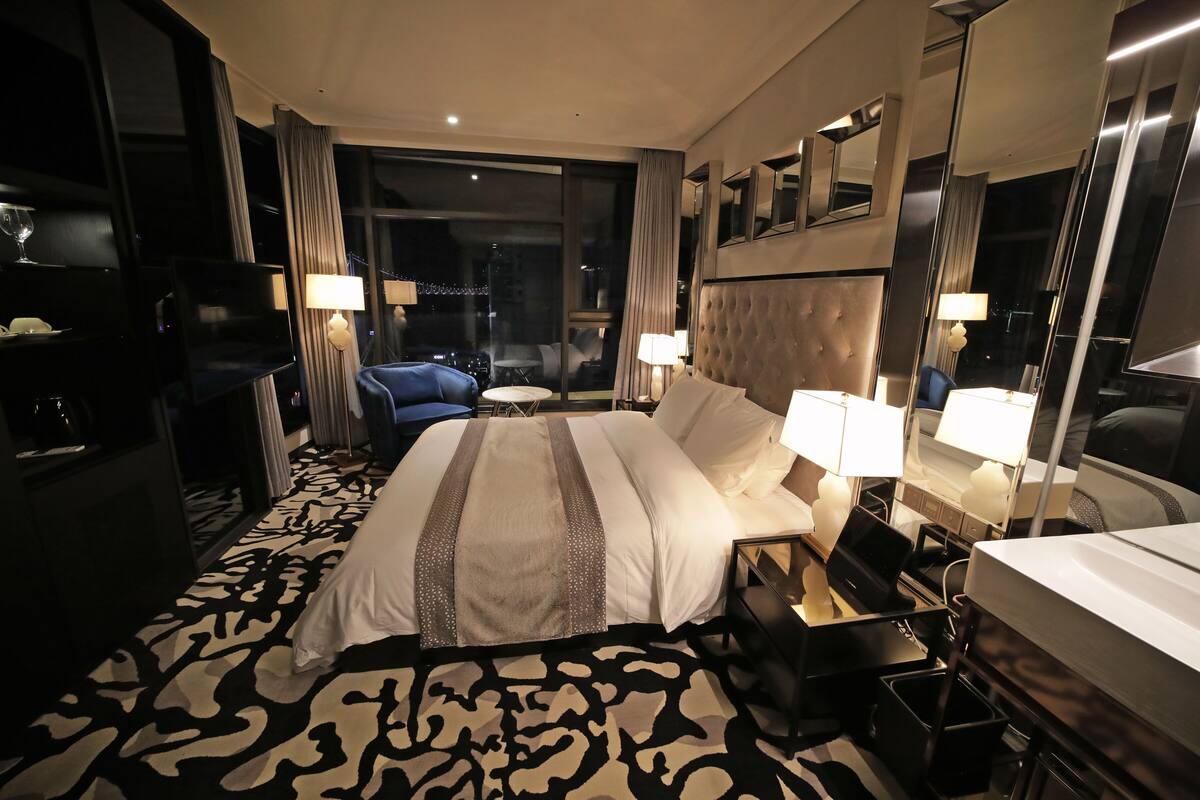 A hotel room with a large bed, a desk with a laptop, a chair, a TV, and a view of the city at night.