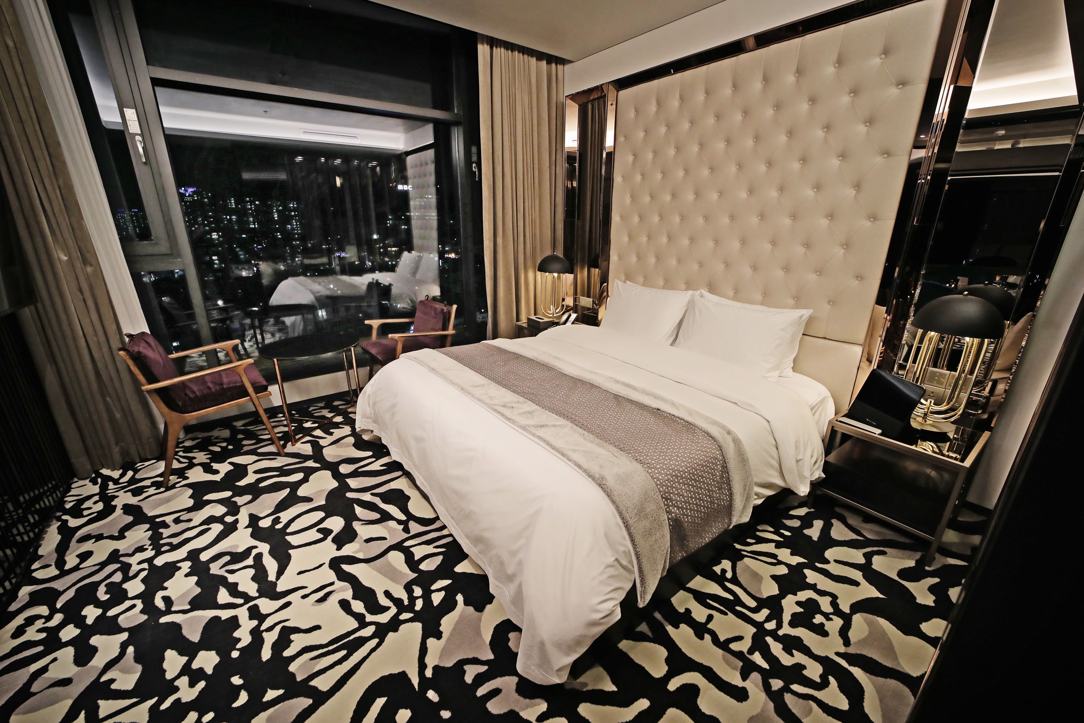 Une chambre d’hôtel avec un grand lit, une vue sur la ville grâce à de grandes fenêtres, un fauteuil et une table de chevet avec une lampe.