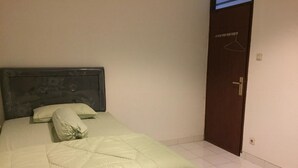 A neatly made bed with a light-colored duvet and pillows, a dark headboard, and a plain wall with a door to the right. - Rumah Kost (Jakarta)
