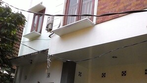 A two-story building with a white facade and a brick section on the upper floor. There are multiple windows, including a balcony with a metal railing. The building has a flat roof and a visible number '9/6' on the lower facade. - Rumah Kost (Jakarta)