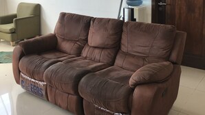 A brown reclining sofa in a living room with a wooden door and a green armchair in the background. - Rumah Kost (Jakarta)