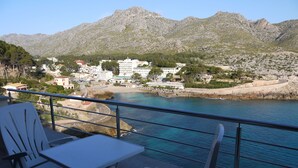 Une terrasse avec des chaises blanches et une table, d’où l’on a vue sur une zone côtière, avec des bâtiments et des montagnes en arrière-plan.