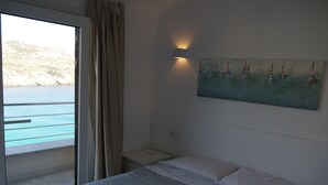 Une chambre d’hôtel avec un lit, une table de chevet, un tableau représentant des voiliers, une fenêtre avec des rideaux et une vue sur la mer.