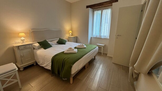 Un dormitorio ordenado con cama, mesitas de noche, un banco y una ventana con cortinas.