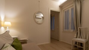 Un dormitorio con una cama, una mesita de noche con lámpara, un espejo, una ventana con cortinas y una silla.