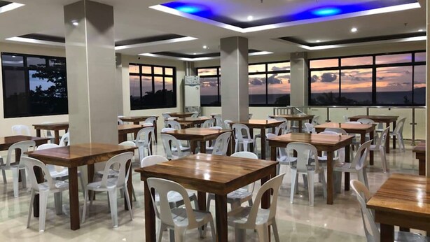 A spacious dining area with multiple tables and chairs, large windows with a sunset view, and modern ceiling lighting. - Godo's Hotel and Restaurant (Culasi)