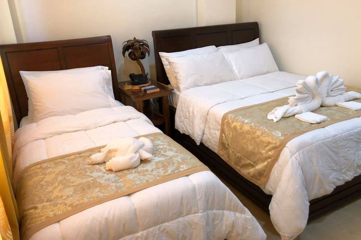 Two adjacent beds with white linens, a wooden headboard, and a small table with a lamp and books between them.