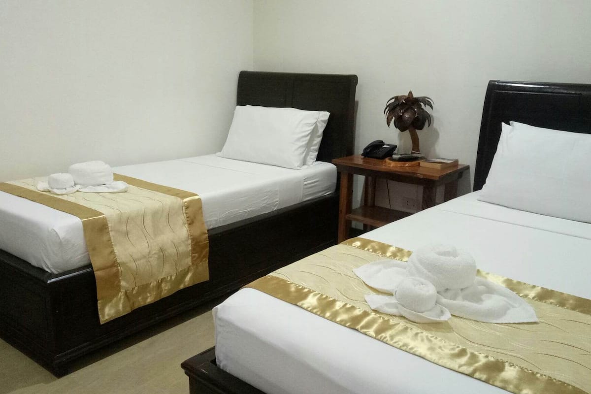 Two single beds with white linens and gold accents, a wooden nightstand with a phone and a small plant, and a plain white wall.