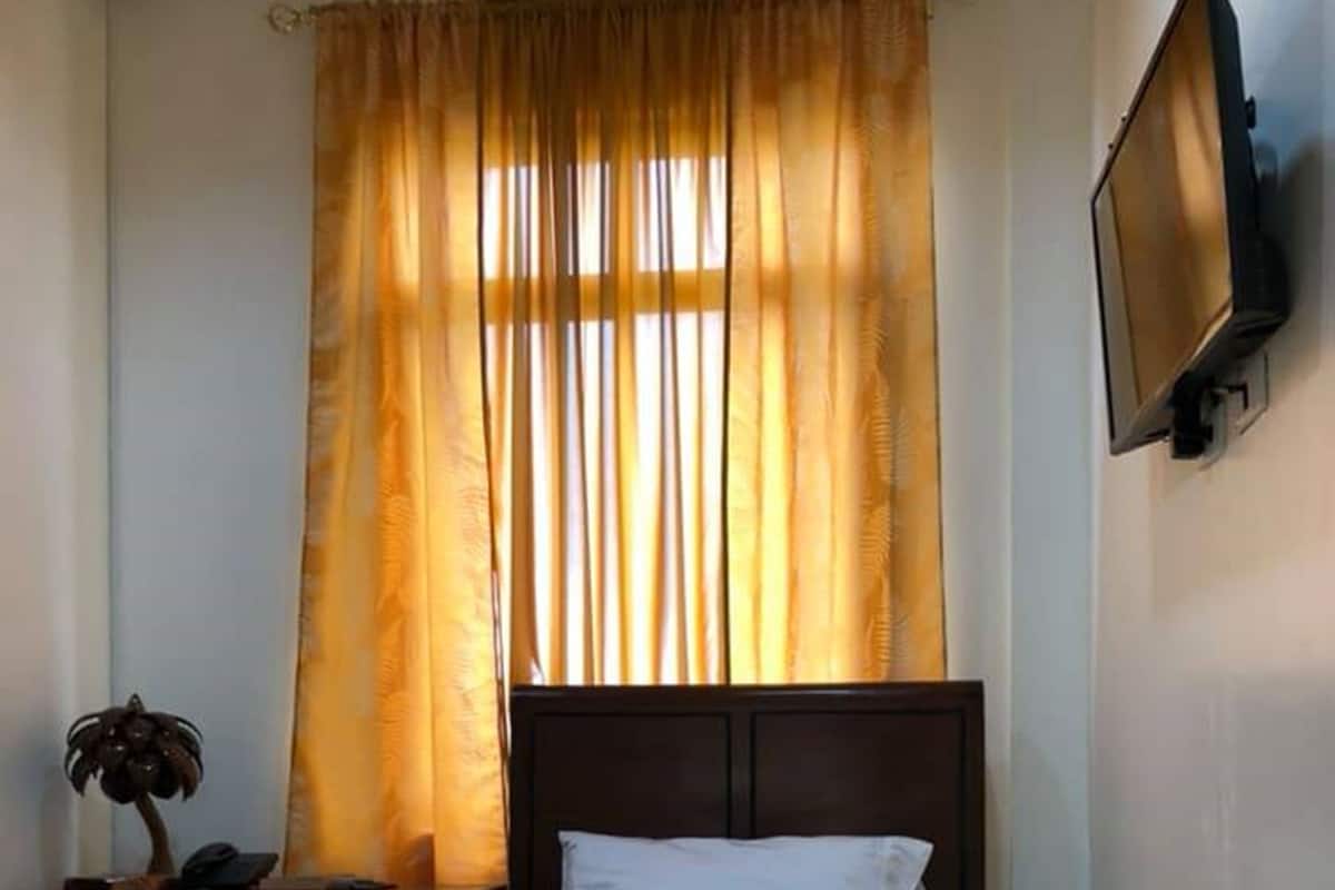 A neatly made bed with white linens and a decorative throw, a wooden nightstand with a lamp, and a television mounted on the wall.
