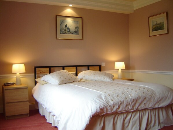 Una cama bien tendida con sábanas blancas y cabecera de madera, flanqueada por dos mesitas de noche con lámparas, en una habitación con paredes color crema y obras de arte enmarcadas sobre la cama.