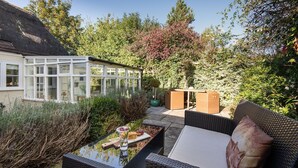 En terrasse med fletmøbler, et glasbord og udsigt til et hus med orangeri.