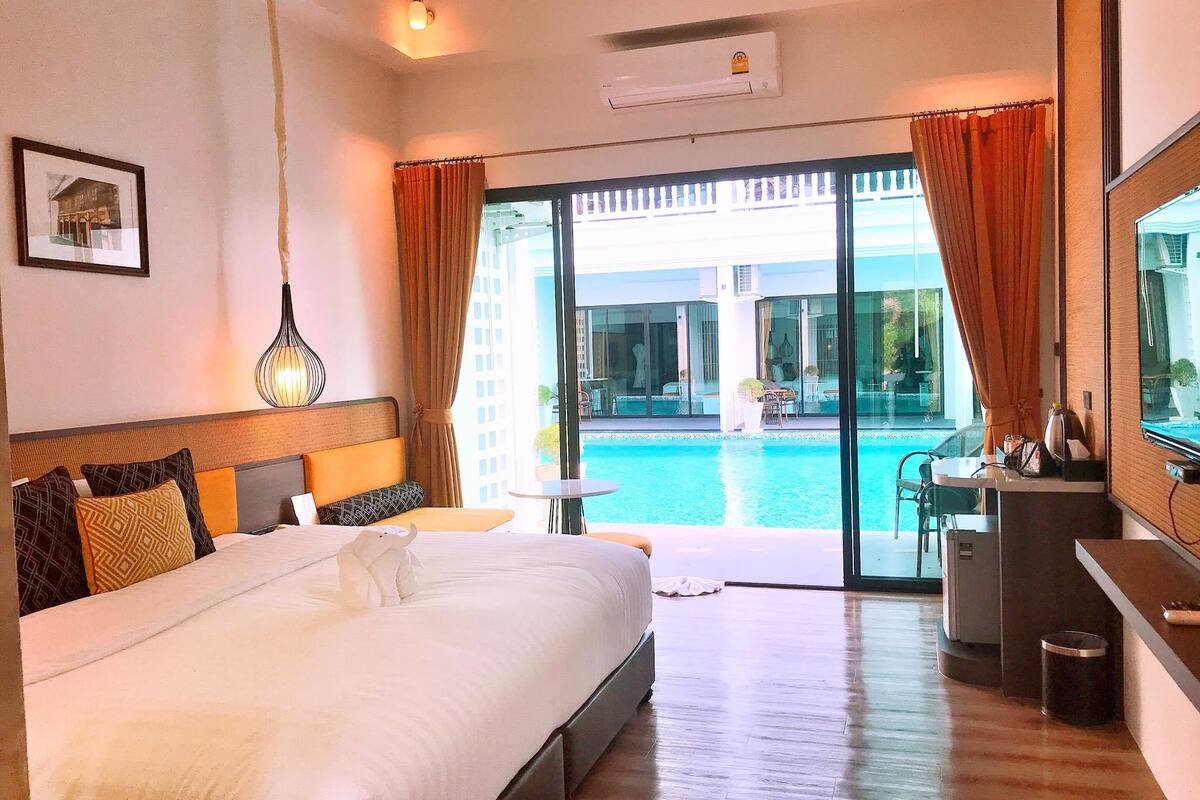 A hotel room with a large bed, a desk, a chair, a small table, a TV, and a view of a swimming pool.