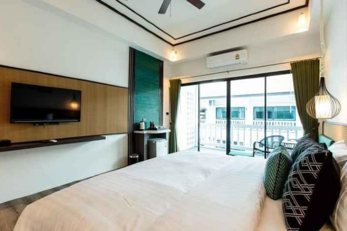 Una habitación de hotel con una cama, televisión montada en la pared, ventana con vista a edificios y un ventilador de techo.