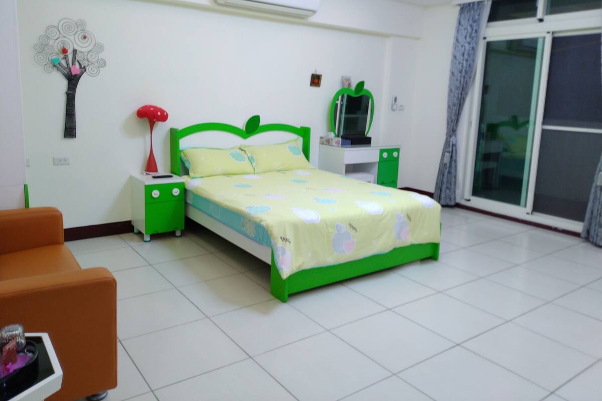Un dormitorio con una cama verde, paredes blancas, una ventana con cortinas, un escritorio y una silla.
