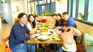 A group of people seated around a wooden table with plates of food, in a room with a large window and a clock on the wall.