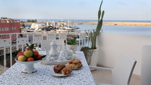 Un balcón con una mesa dispuesta para el desayuno, con vistas a un puerto deportivo con barcos y un mar tranquilo.