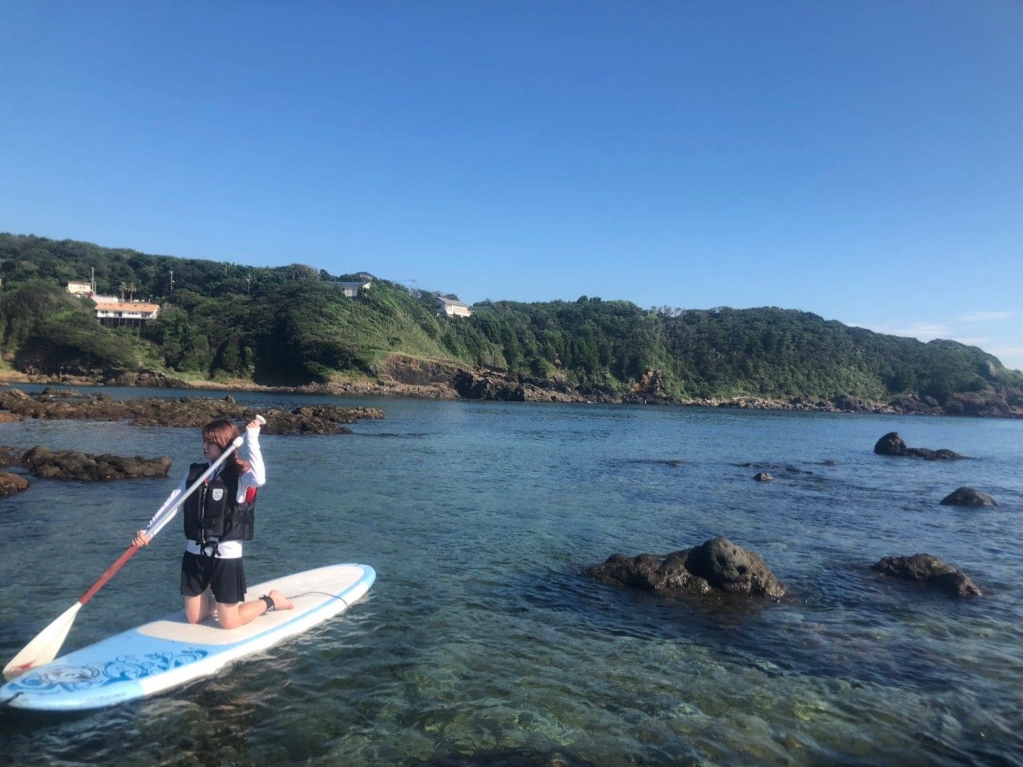 A person standing on a paddleboard in clear water with rocks, a green hillside in the background, and houses on the hill.