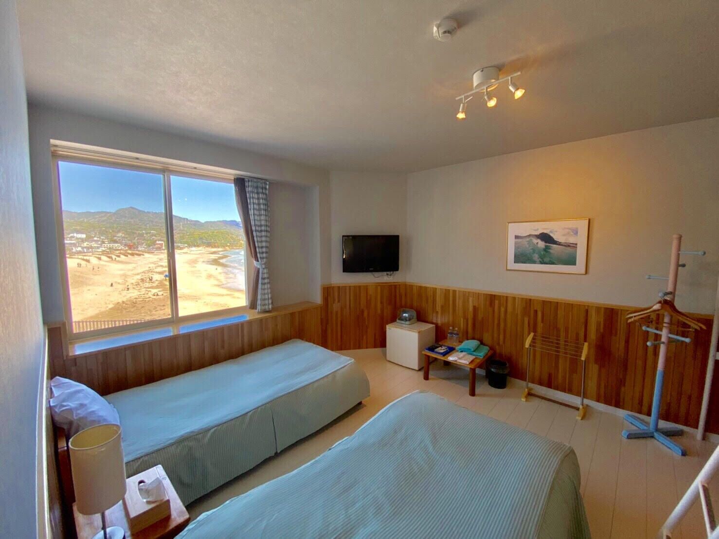 A room with two beds, a TV, a painting, and a window with a view of a beach.