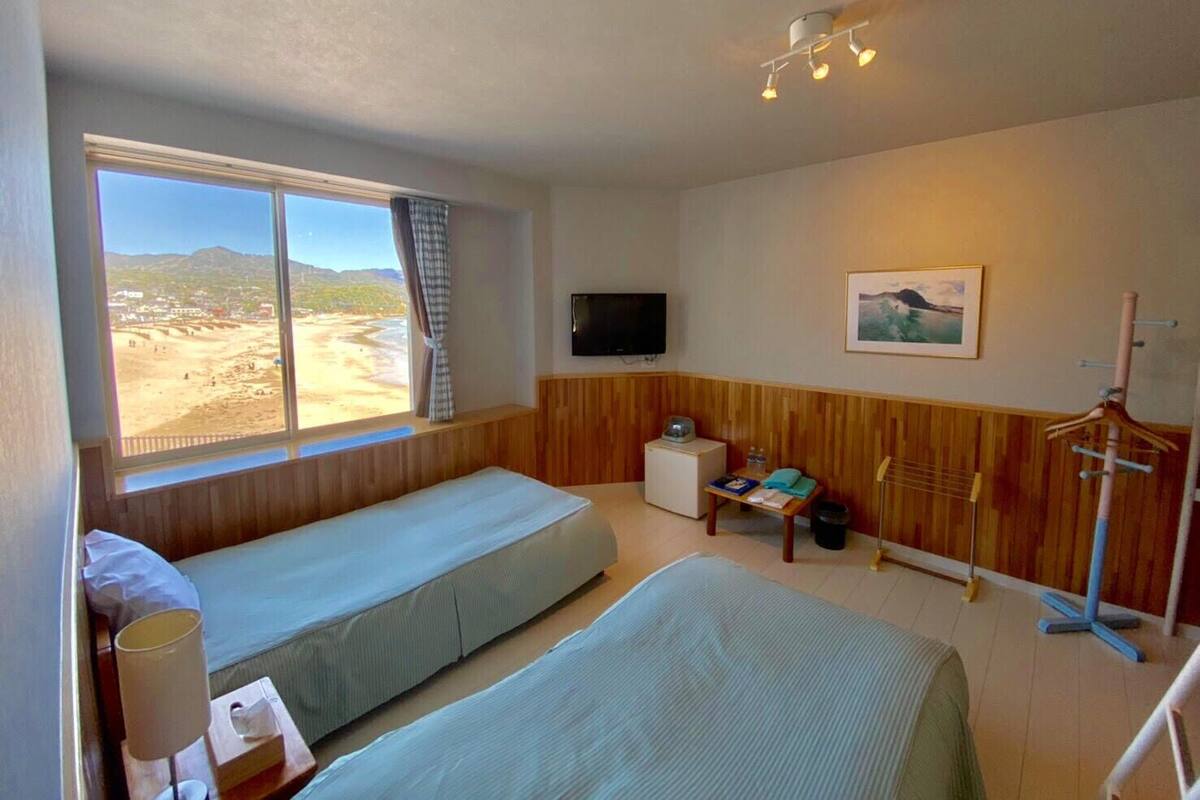 A room with two beds, a TV, a painting, and a window with a view of a beach.