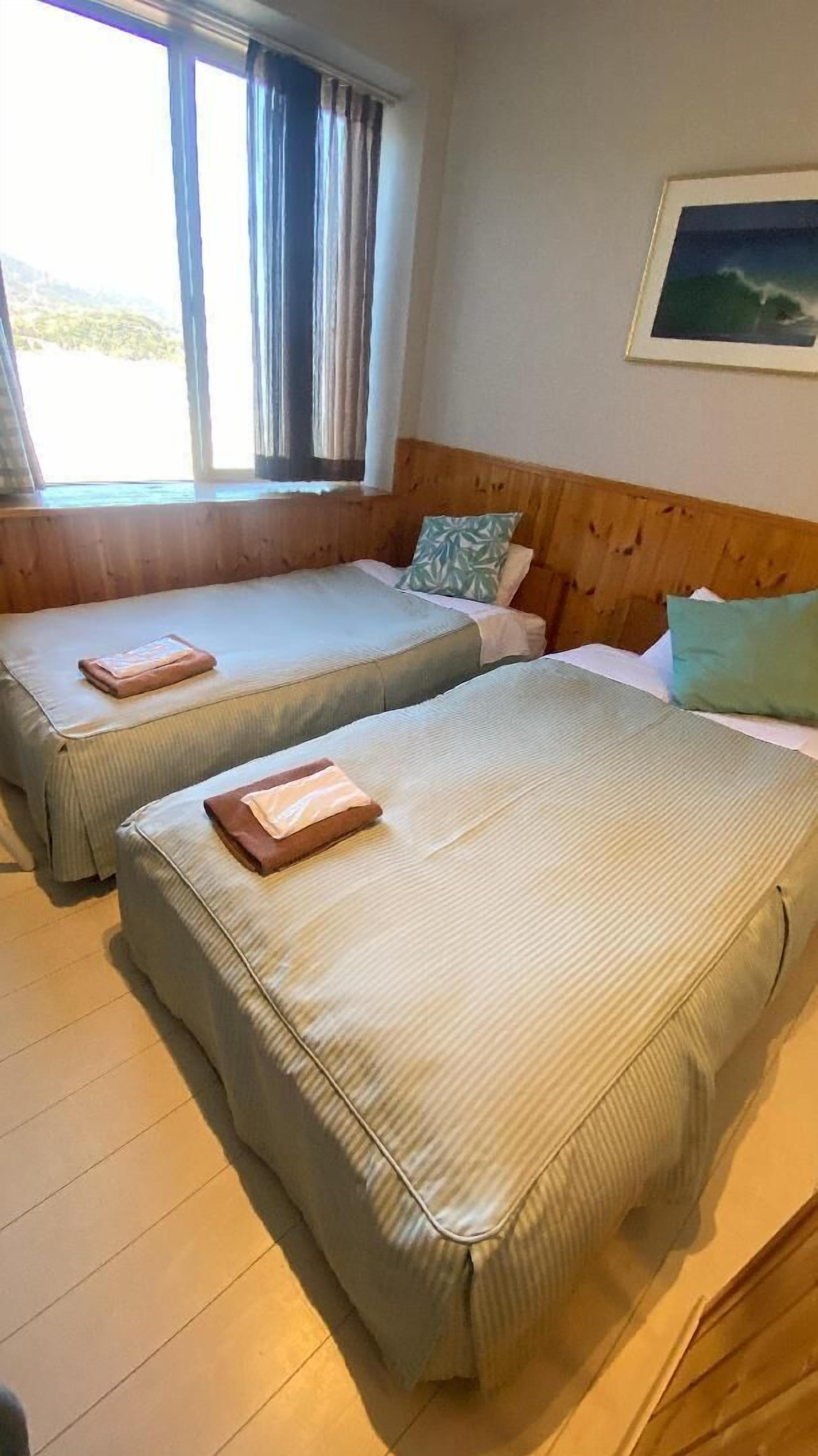 Two single beds with pillows and folded towels, a window with curtains, a framed picture on the wall, and a wooden headboard.
