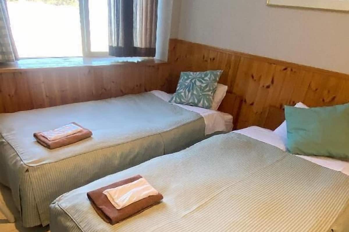 Two single beds with pillows and folded towels, a window with curtains, a framed picture on the wall, and a wooden headboard.