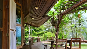 Una terraza de madera con mesas y sillas, rodeada de exuberante vegetación y árboles.