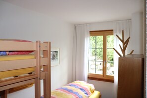 Una habitación con una cama litera, una cama individual con un edredón colorido, un ventanal con cortinas y un armario de madera.