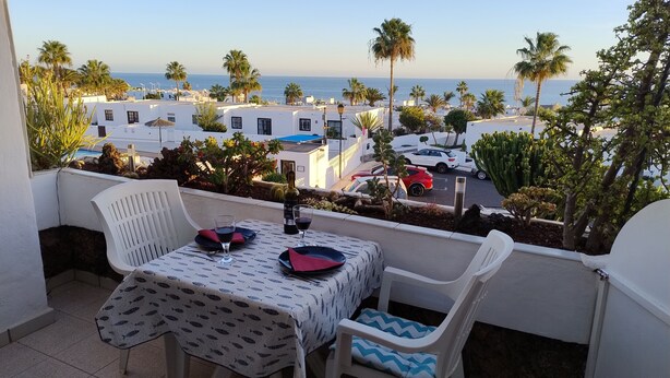 Un balcón con una mesa para dos, con vista a una zona costera con palmeras y edificios blancos.