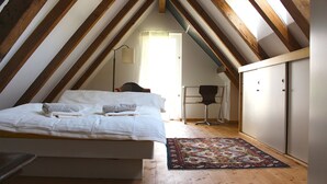 Une chambre avec un plafond en pente, un lit recouvert de draps blancs, un tapis à motifs et une chaise.