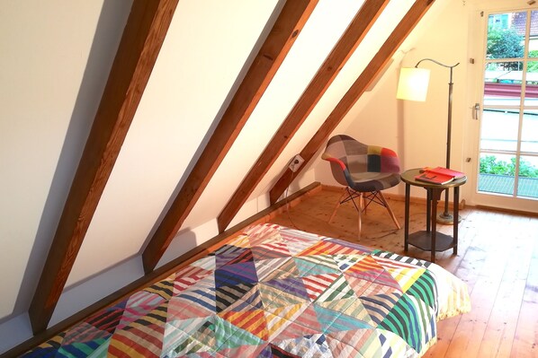 A room with a sloped ceiling, a colorful quilt on the bed, a chair, a small table, and a lamp.